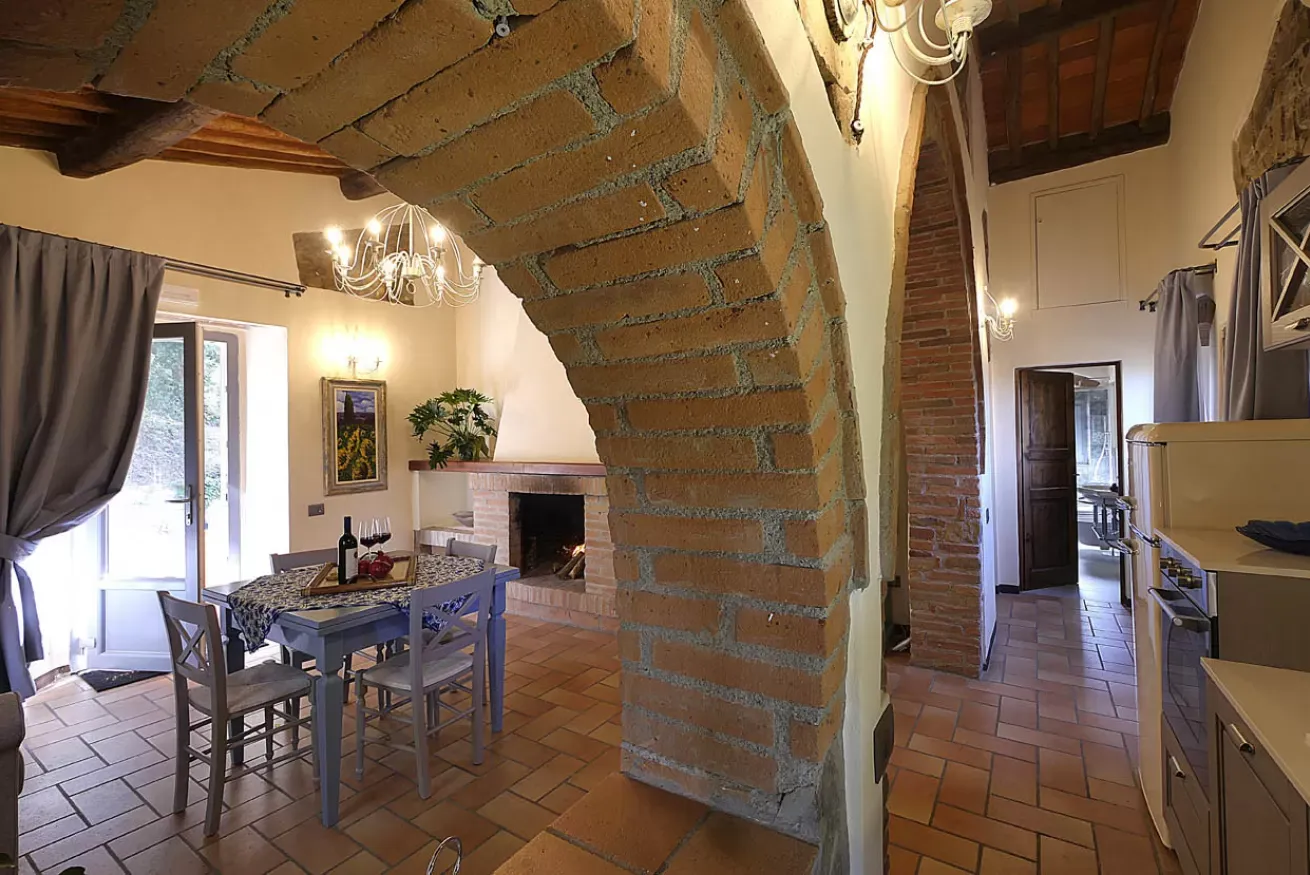 Interno rustico con arco in mattoni, tavolo da pranzo, camino e cucina sullo sfondo