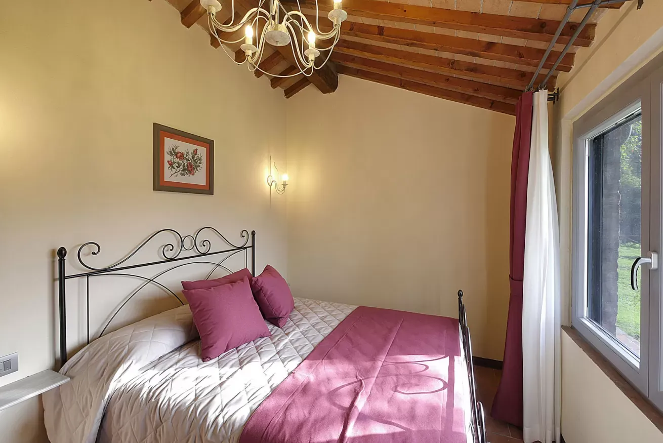 Camera da letto con letto in ferro battuto, cuscini viola, soffitto con travi in legno e finestra laterale