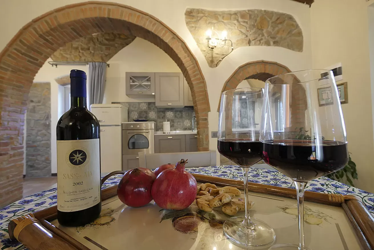 Tavola con bottiglia di vino, due calici, melograni e biscotti in una cucina rustica con archi in pietra