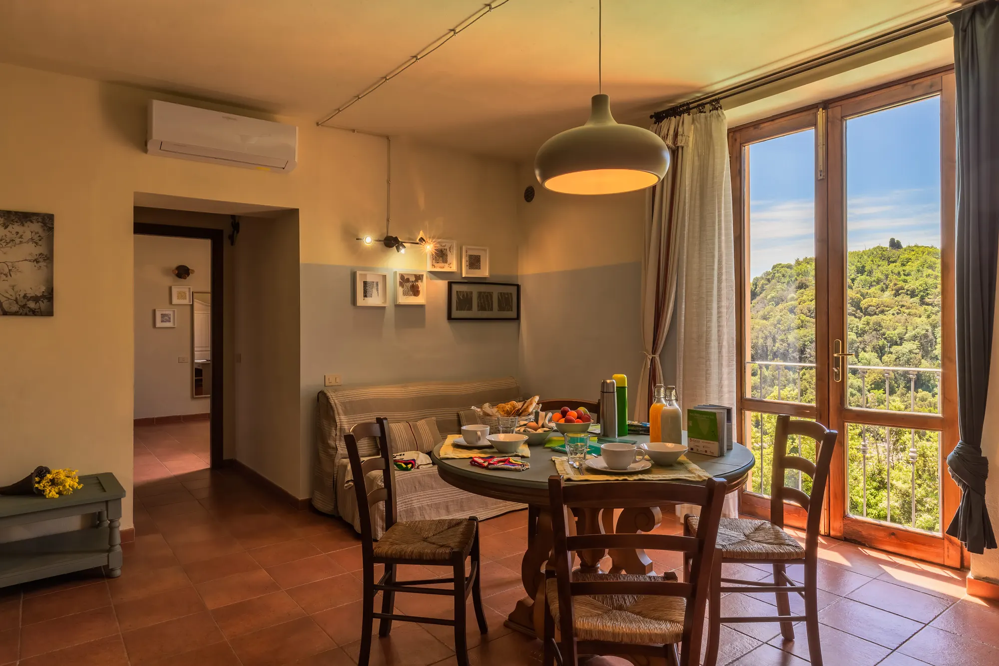 Sala da pranzo con tavola apparecchiata, grande finestra aperta e vista su colline verdi