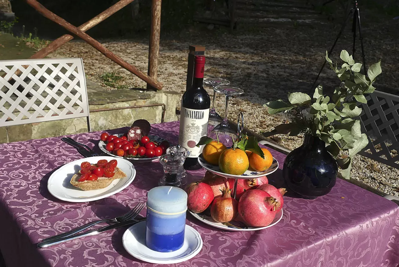 Tavola all’aperto con frutta, vino, bicchieri e un vaso di foglie su una tovaglia viola