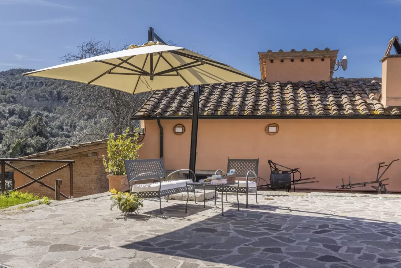 Terrazza esterna con tavolino, sedie e grande ombrellone accanto a una casa con tetto in tegole