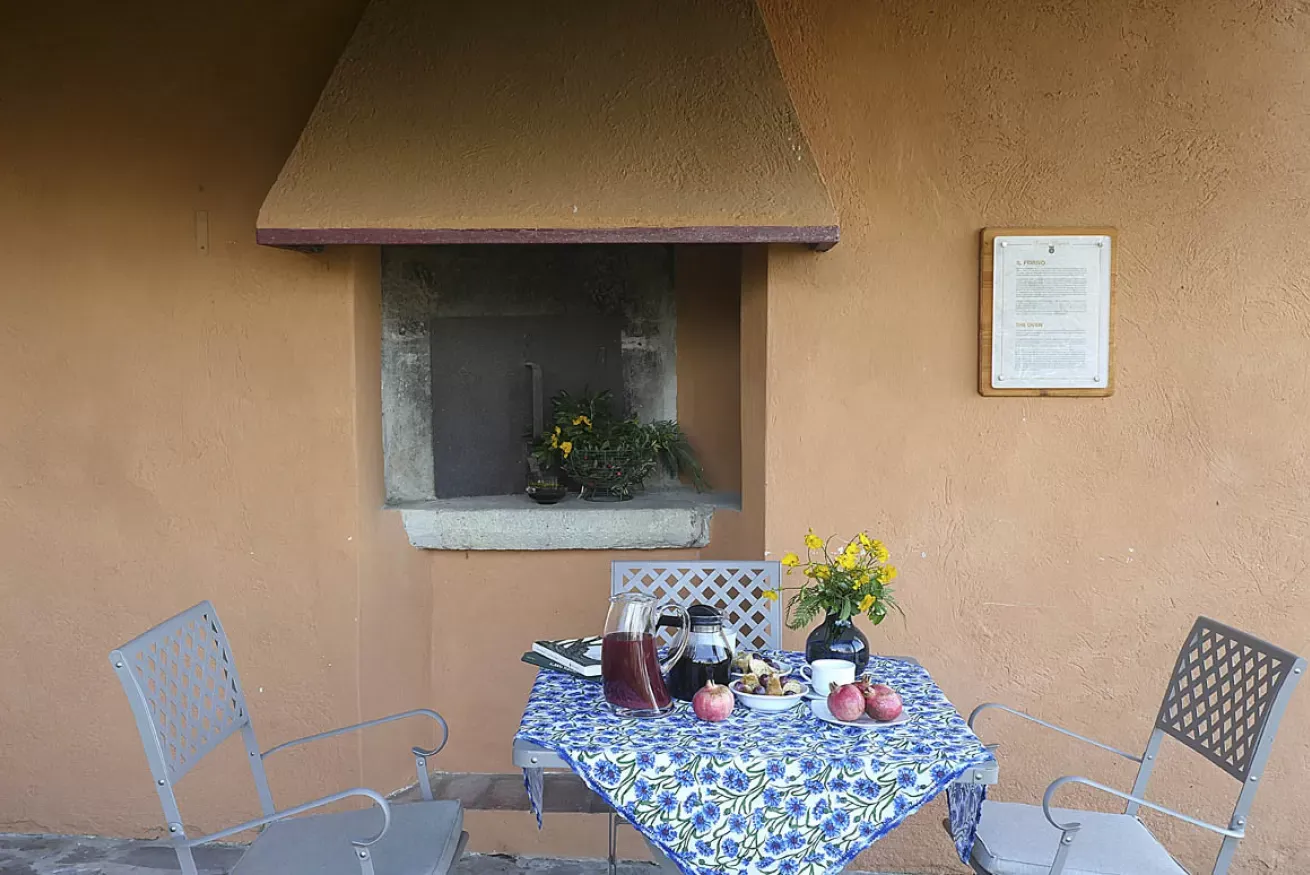 Tavolo apparecchiato all’aperto con brocca, frutta e fiori, davanti a una parete color ocra