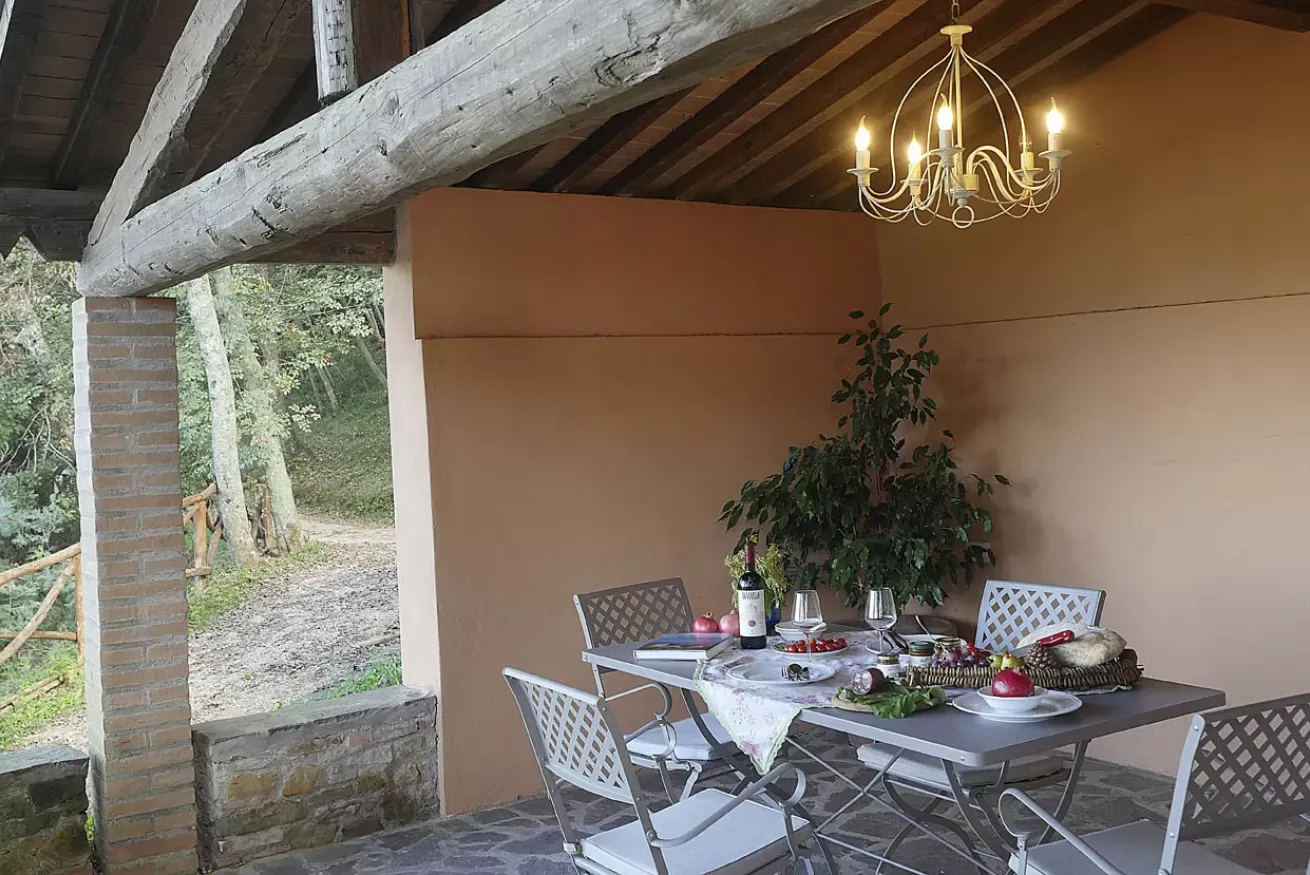 Tavolo apparecchiato sotto un portico rustico con lampadario acceso e vista sul bosco