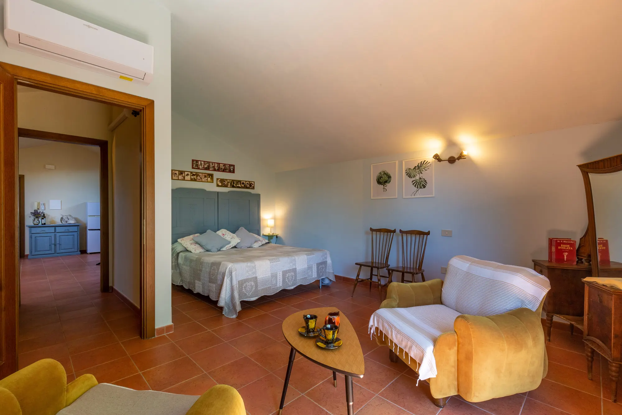 Camera da letto con letto matrimoniale, poltrona, tavolino e due sedie, vista su un angolo con mobile e frigorifero