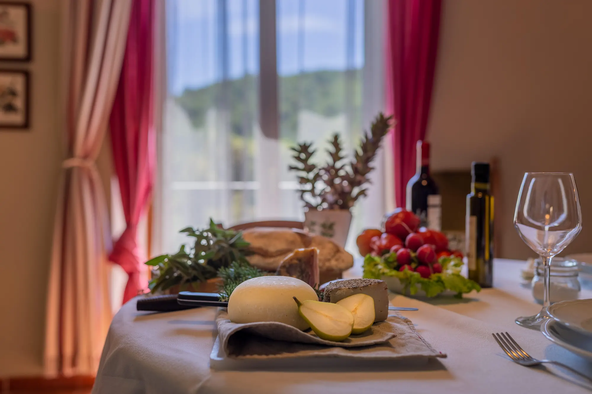 Tavola apparecchiata con formaggi, pane, frutta e vino davanti a una finestra