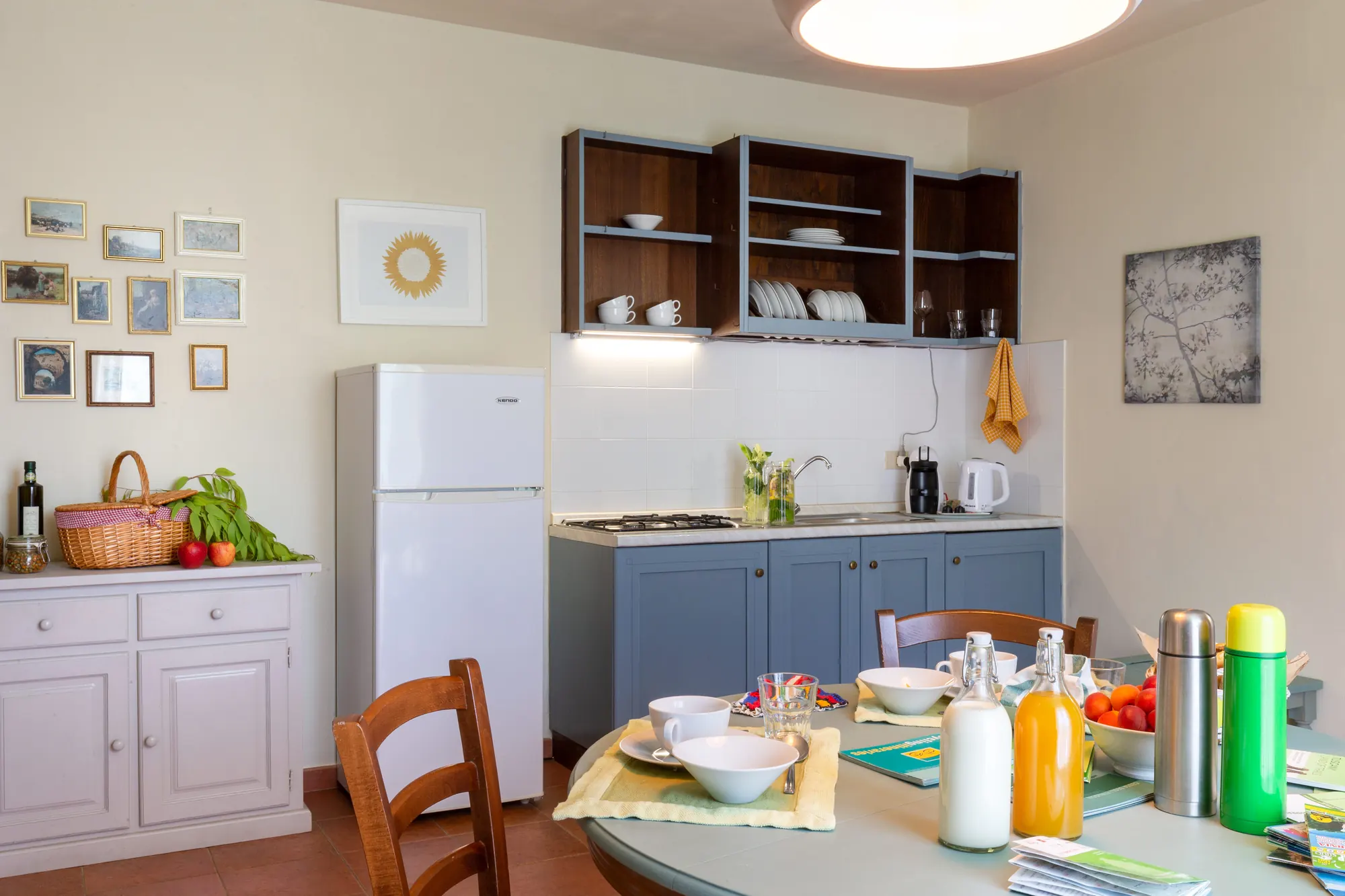Cucina luminosa con tavolo apparecchiato, frigorifero bianco e mobili blu con stoviglie a vista