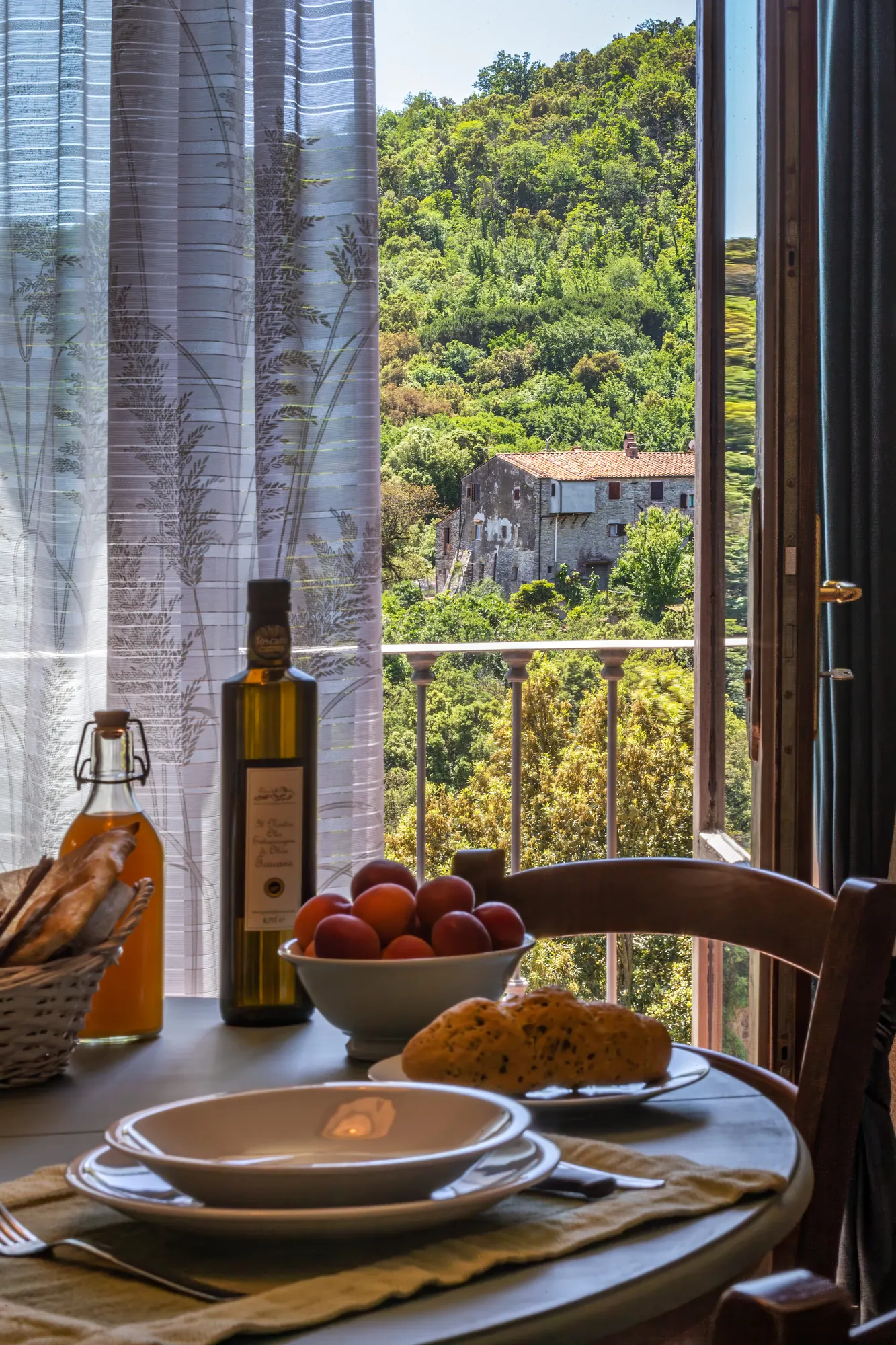 Tavola apparecchiata accanto a una finestra aperta con vista su colline verdi e una casa in pietra