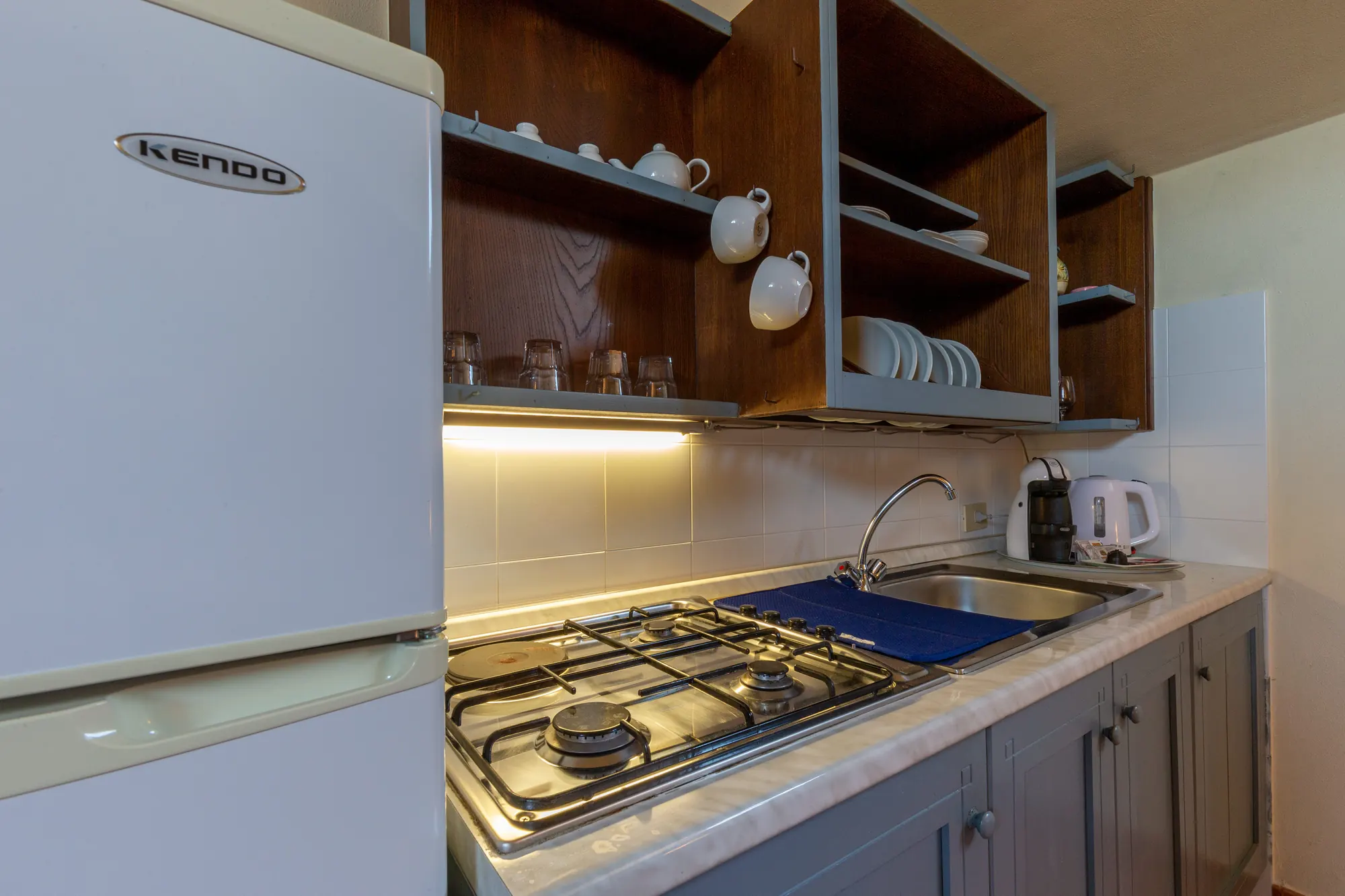 Cucina con frigorifero, piano cottura, lavello e pensili aperti con tazze e piatti