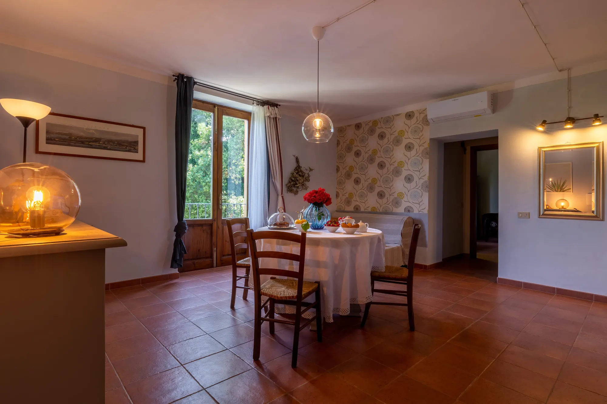 Sala da pranzo luminosa con tavolo apparecchiato, sedie, finestra aperta e lampade accese