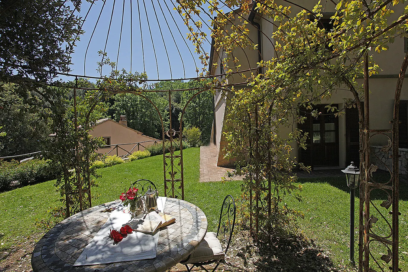Giardino con tavolo rotondo apparecchiato sotto un pergolato, accanto a una casa