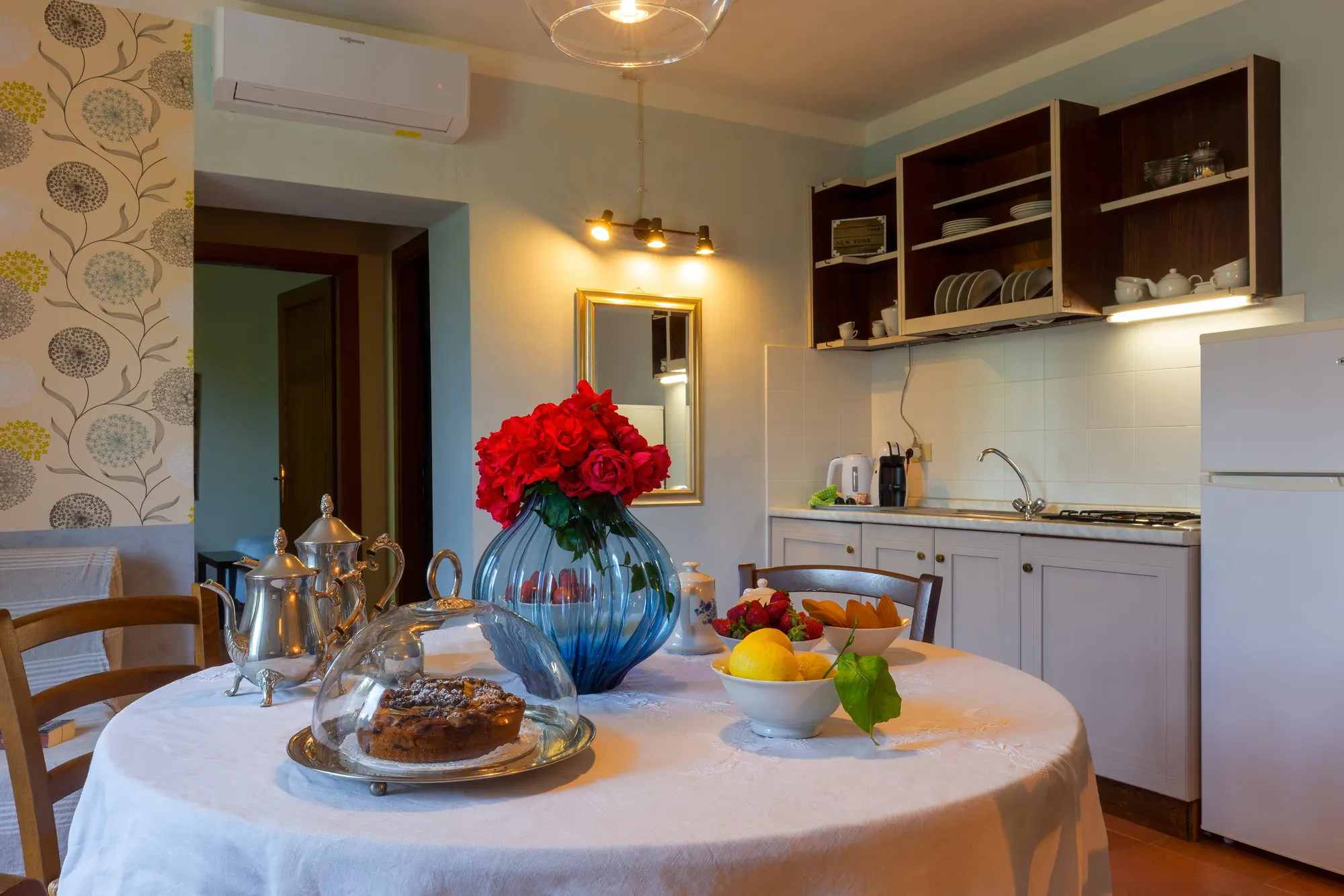 Cucina con tavolo apparecchiato, vassoio con dolce, frutta e vaso di rose rosse al centro
