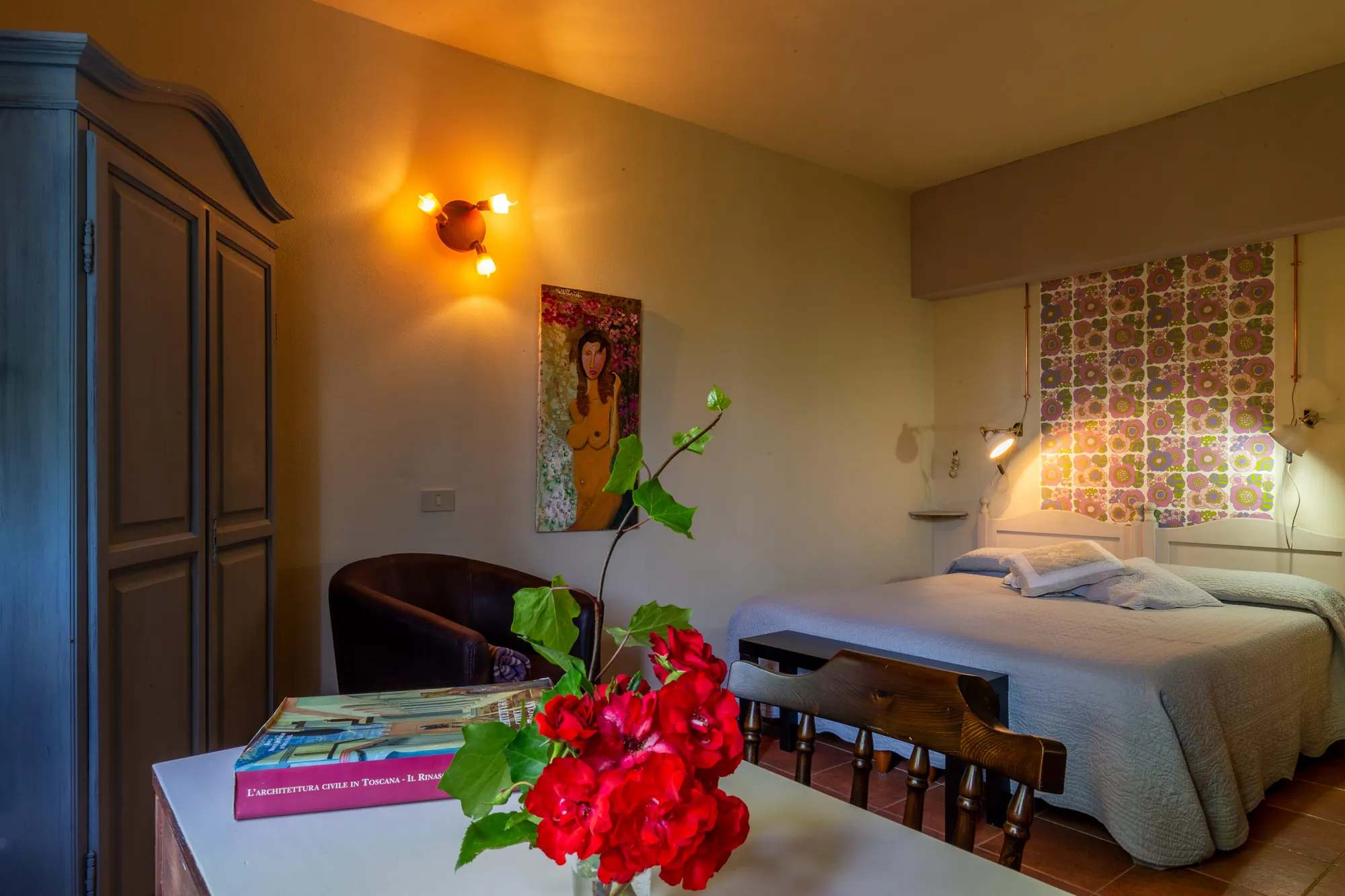Camera da letto con letto matrimoniale, lampade accese e tavolo con fiori rossi in primo piano