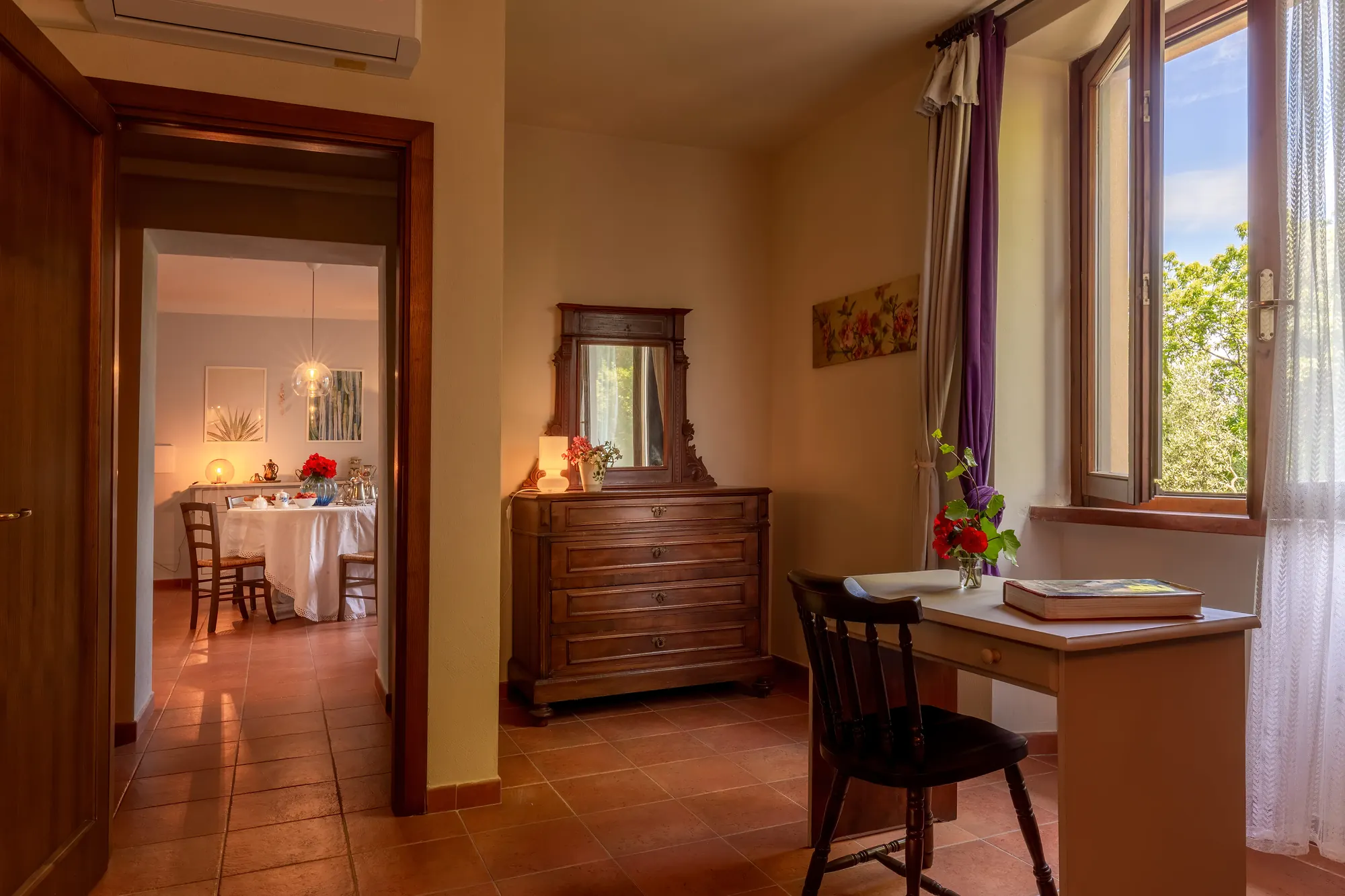 Interno luminoso con scrivania, cassettiera antica e finestra aperta; sullo sfondo una sala da pranzo appare attraverso la porta