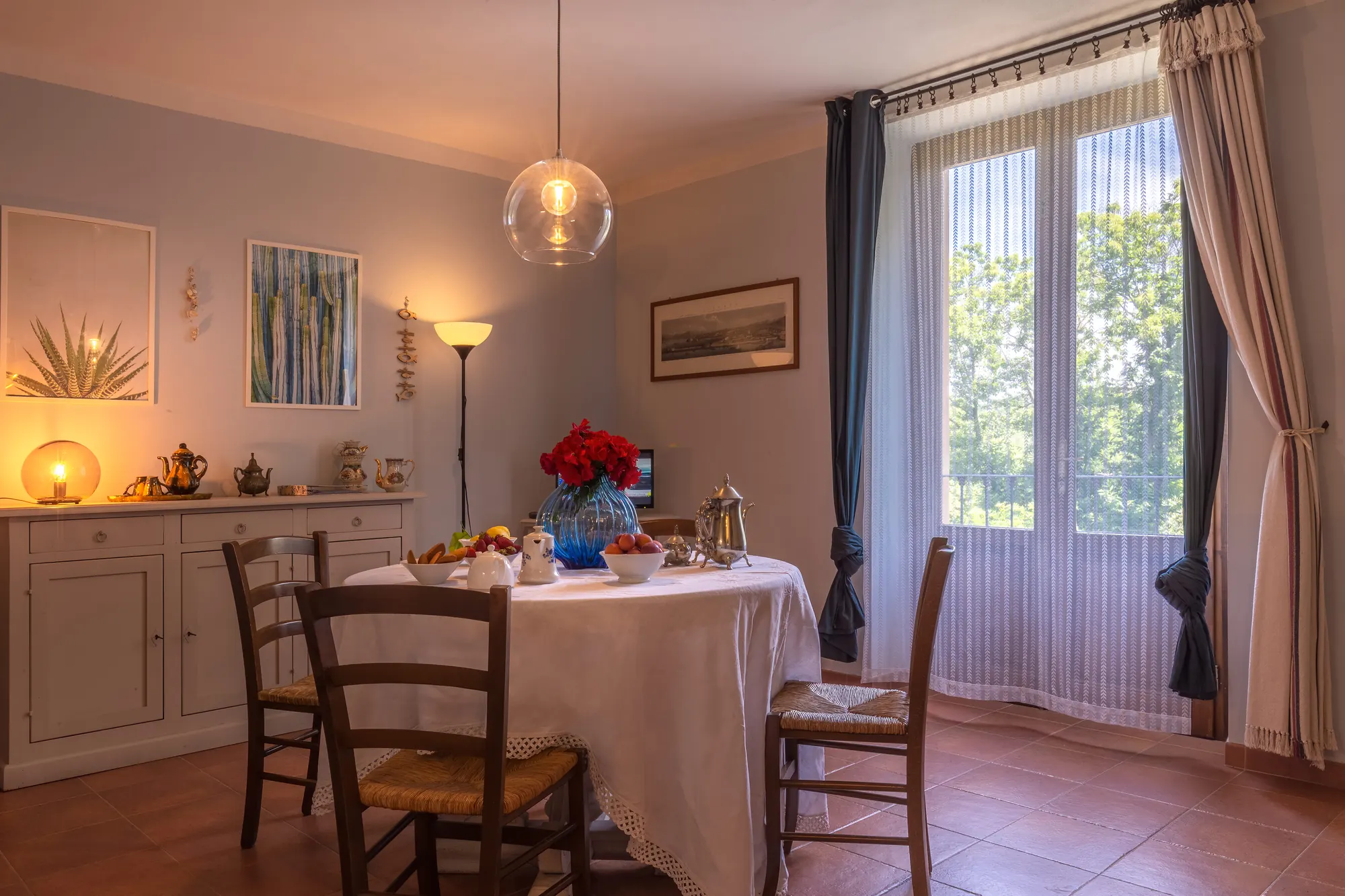Sala da pranzo luminosa con tavolo apparecchiato, sedie, lampada a sospensione e grande finestra con tende