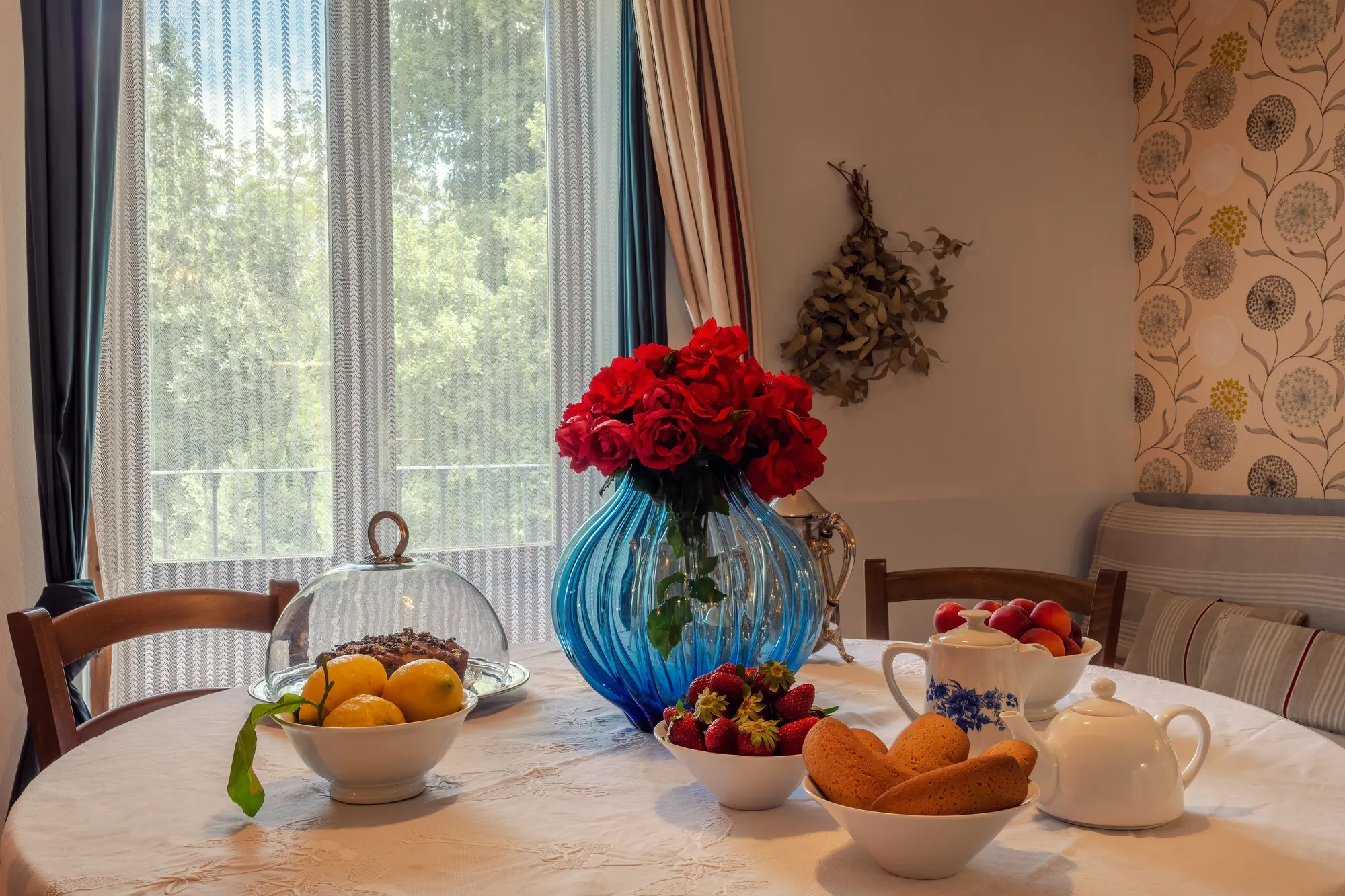 Tavola apparecchiata con frutta, dolci, tè e un vaso blu con rose rosse vicino alla finestra