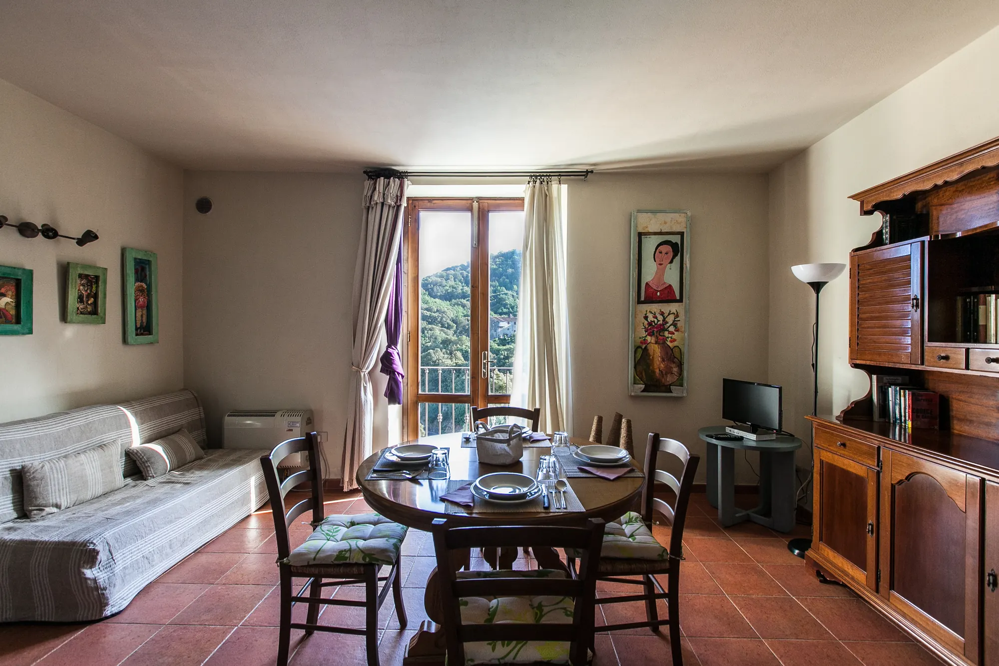 Soggiorno con tavolo apparecchiato, divano, mobile in legno e finestra con vista sulle colline