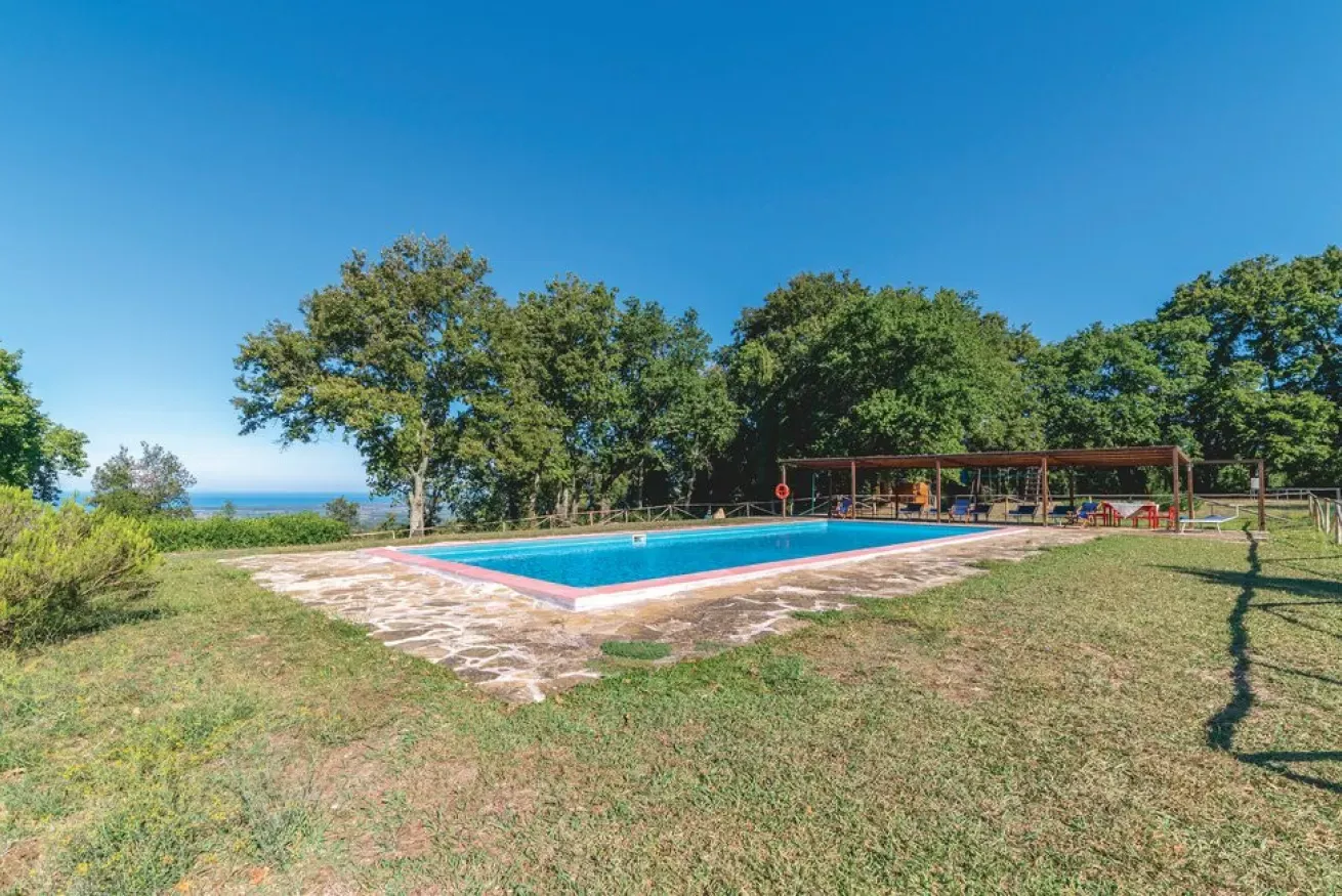 Outdoor swimming pool surrounded by grass and trees with a shaded seating area and distant sea view