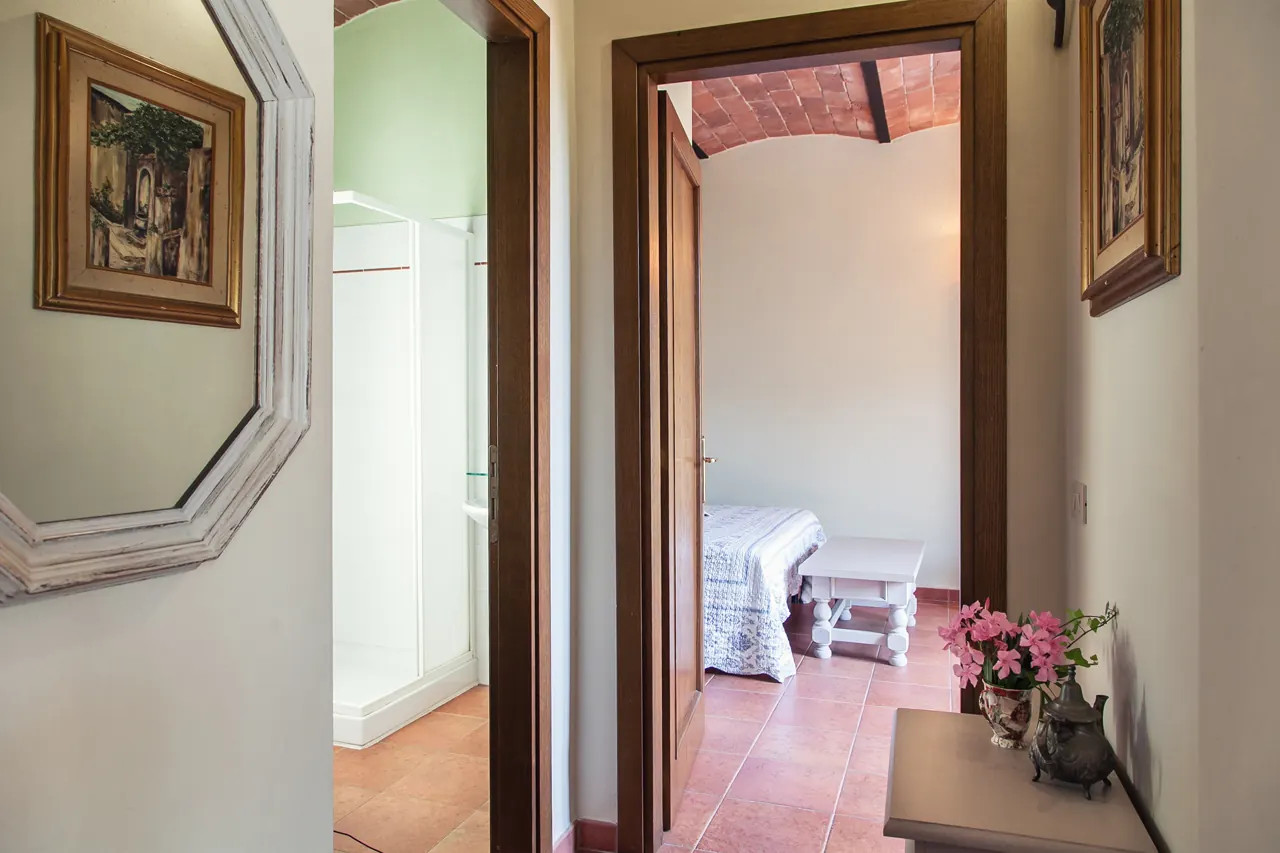 Corridoio interno con porte aperte verso una camera da letto e un bagno, pavimento in cotto e quadri alle pareti