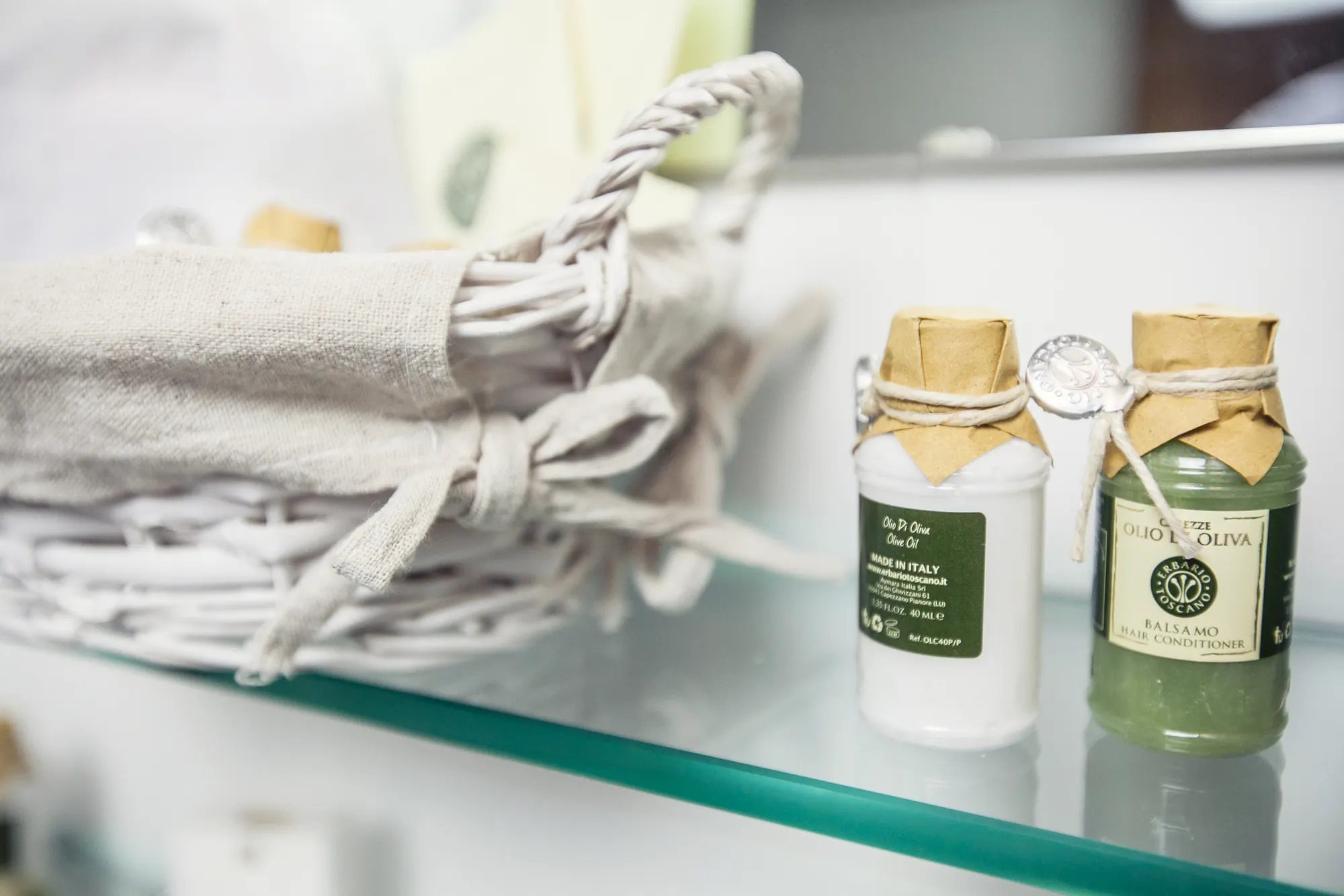 Due flaconi di cosmetici con etichette in italiano su un ripiano di vetro, accanto a un cestino intrecciato