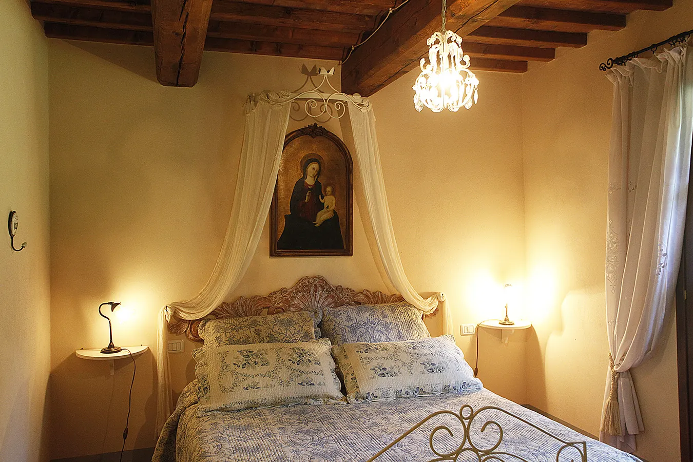 Camera da letto accogliente con letto matrimoniale, lampade accese, travi in legno e quadro della Madonna sopra la testiera