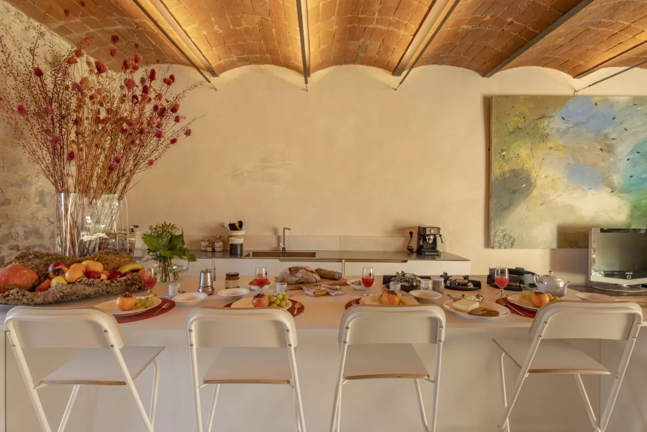 Tavolo apparecchiato per la colazione in una cucina con frutta, pane, bevande e sedie bianche davanti al bancone