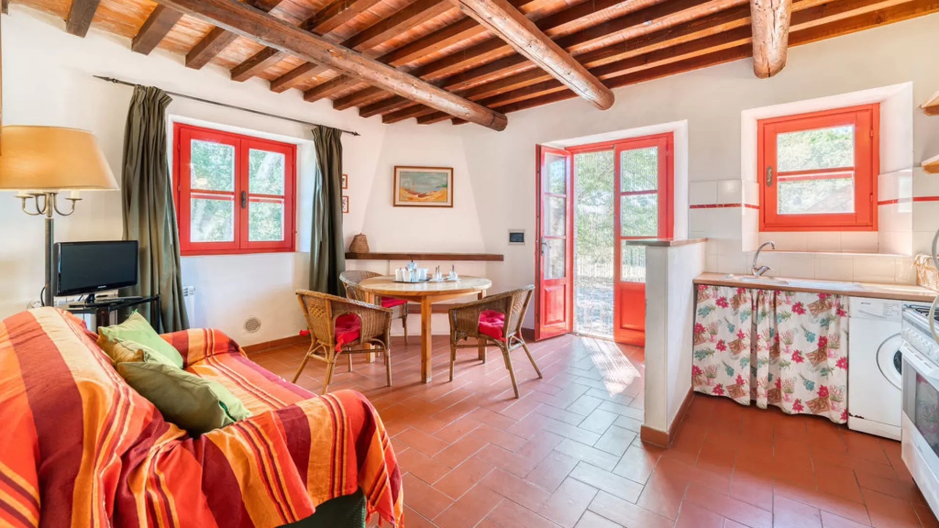 Cozy living-dining area with exposed wooden beams, red-trimmed windows, and a small kitchen corner