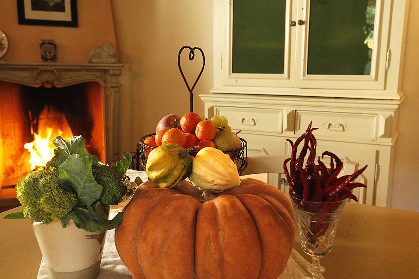 Composizione autunnale con zucca, frutta, broccoli e peperoncini su un tavolo davanti al camino acceso