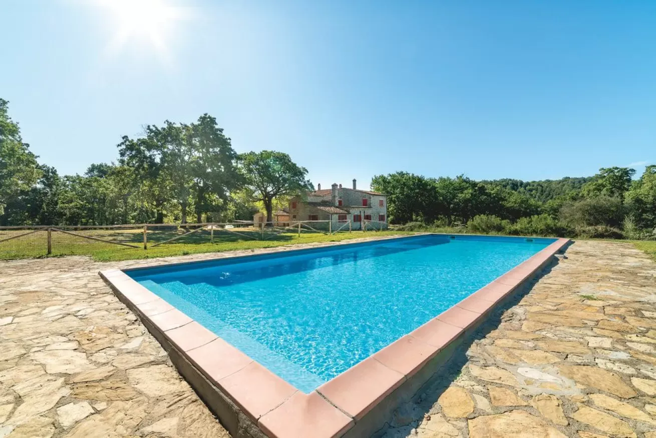 Outdoor swimming pool beside a stone patio with a house and trees in the background