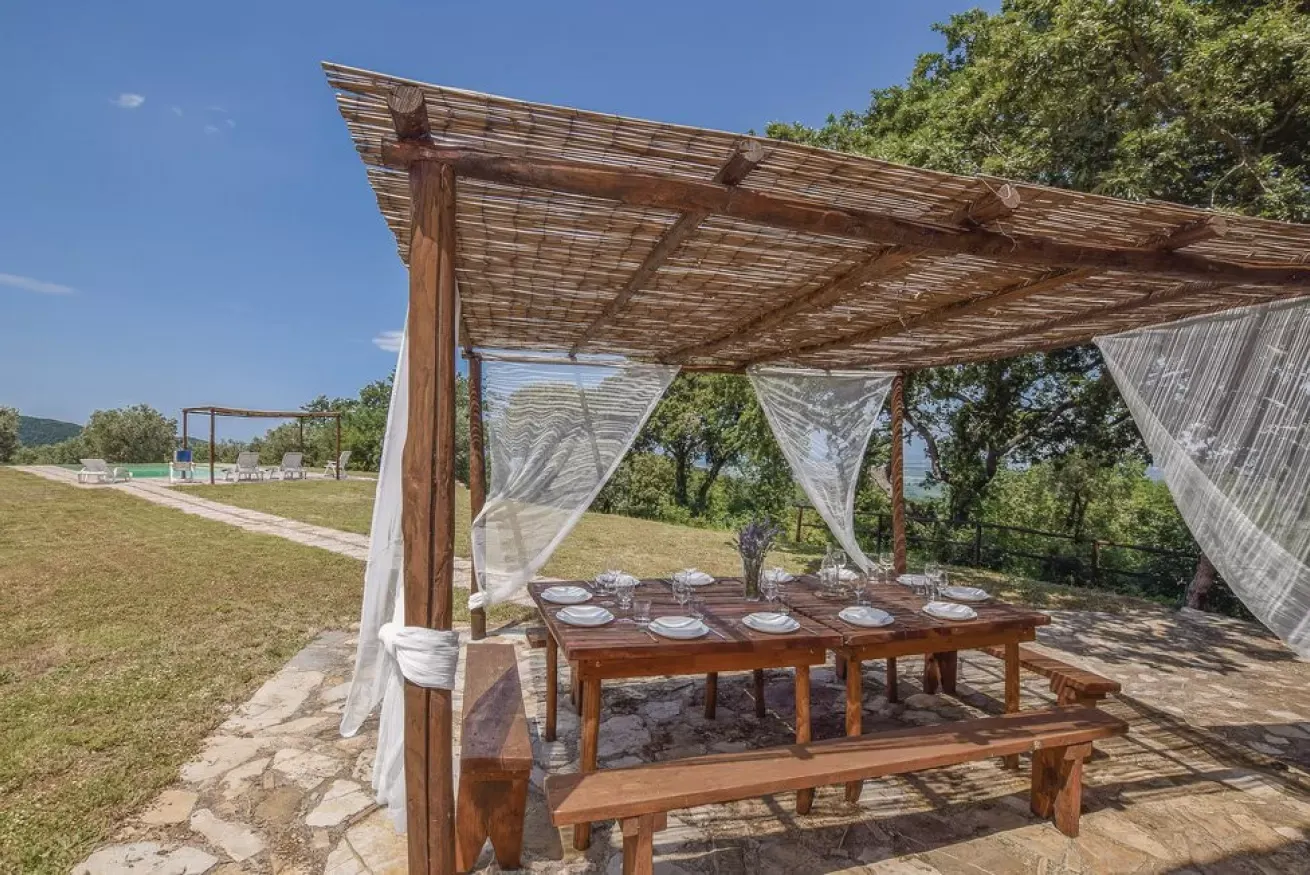Outdoor dining area with a wooden pergola, set table, and sheer curtains overlooking a grassy landscape