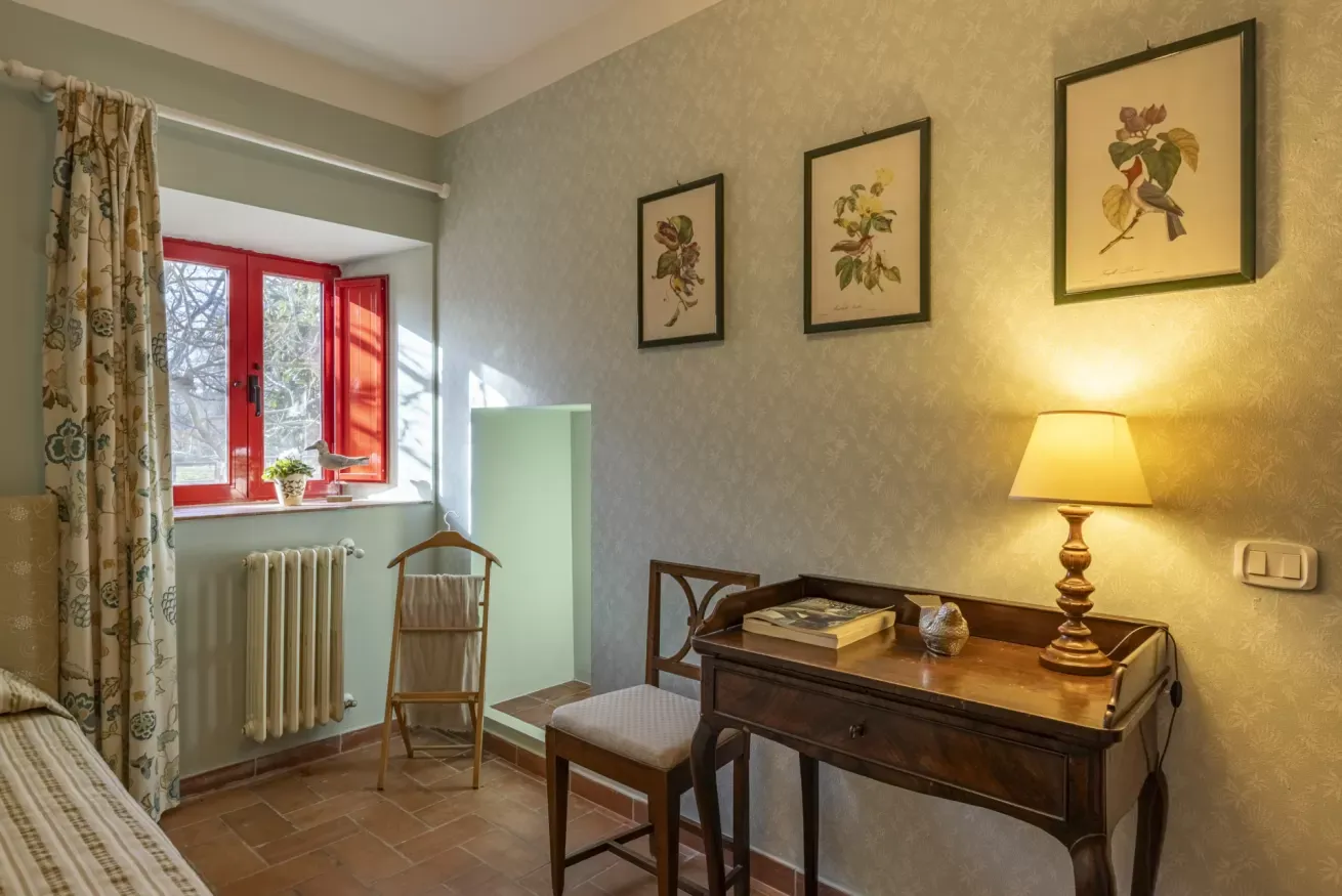 A cozy bedroom with a red-framed window, wooden desk lamp, chair, and framed bird prints on the wall