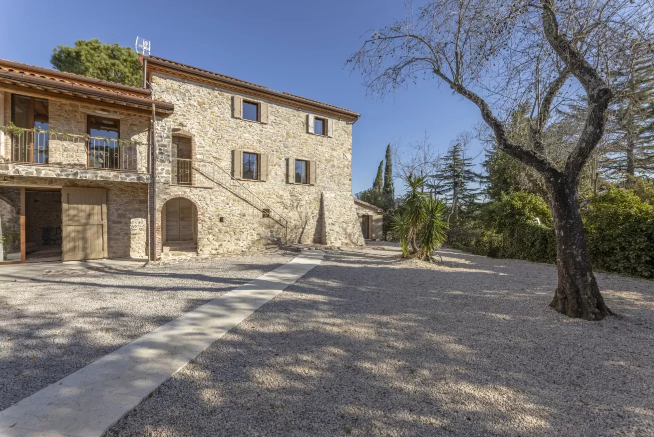 Cortile ghiaioso con casa in pietra e alberi, sotto un cielo sereno