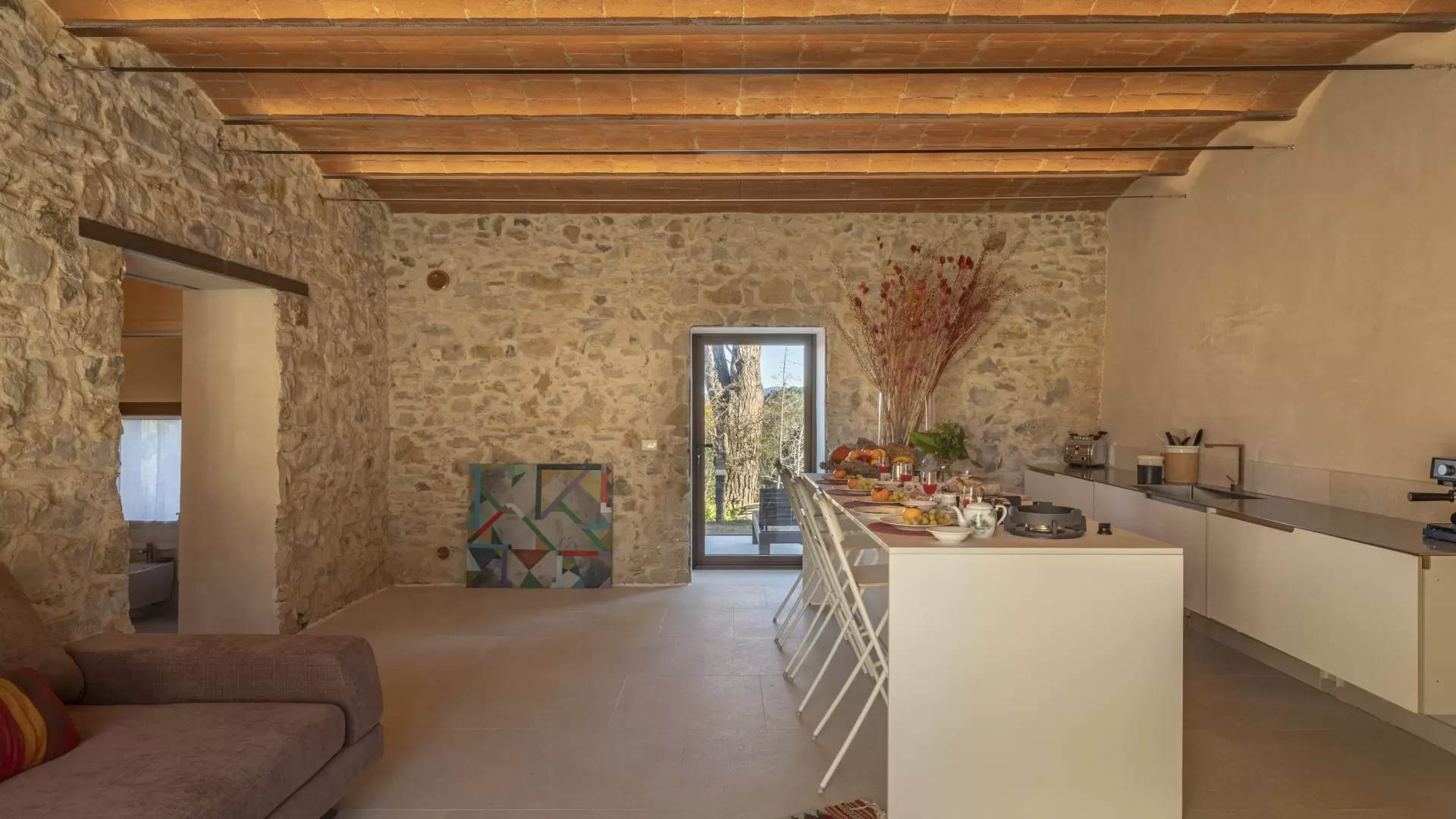 Interno di cucina-soggiorno rustico con pareti in pietra, isola apparecchiata e porta sul giardino