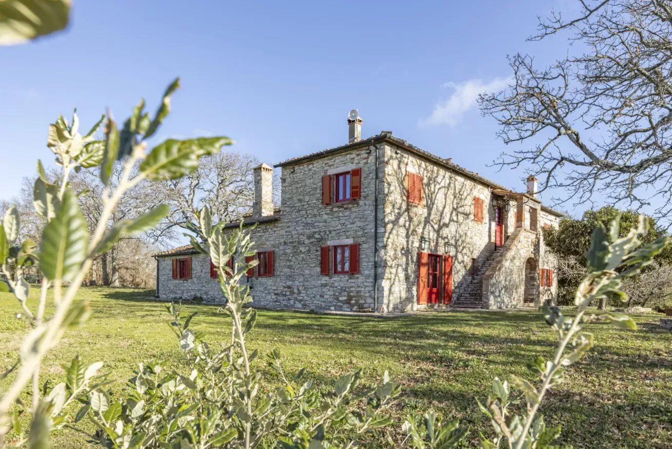 Stone farmhouse with red shutters in a grassy yard, framed by trees and foliage